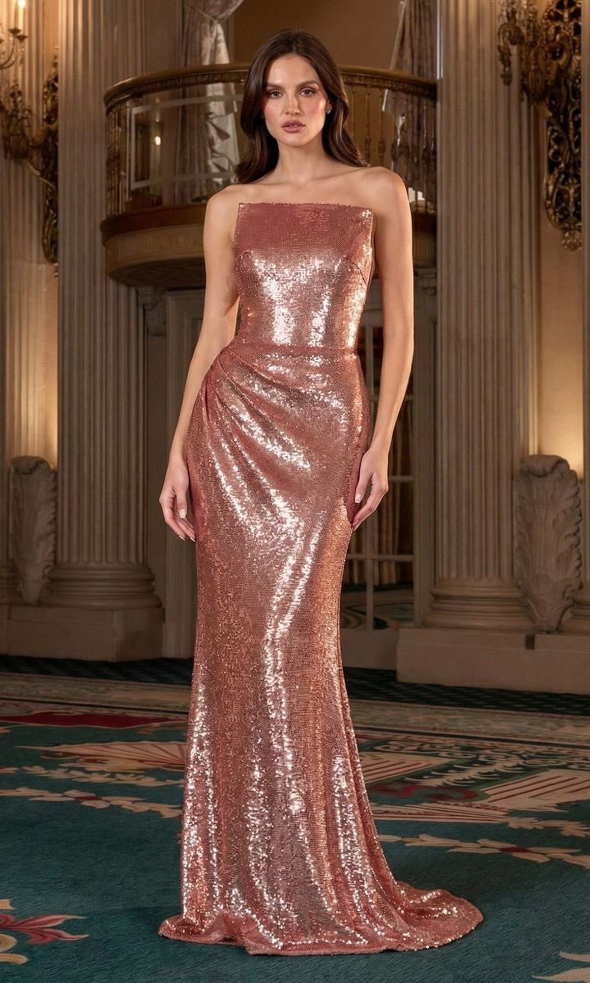 A woman stands indoors wearing the Ladivine J904, a floor-length rose gold prom dress with sequins and a square neckline. Ornate columns and railings can be seen in the background.